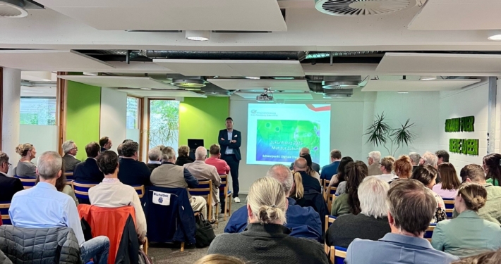 Eine Veranstaltung in einem Konferenzraum. Man sieht die Teilenehmenden mit dem Rücken in Stuhlreihen sitzen.
