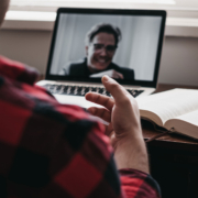 Eine Person wird von hinten angeschnitten gezeigt, die an einem Schreibtisch sitzt auf dem ein Laptop aufgeklappt steht und ein Mann mittleren Alters zu sehen ist. Die Szene spiegelt ein Videokonferenz wider.