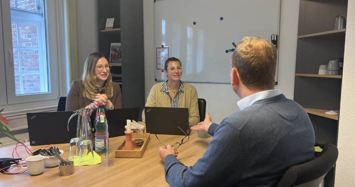 WelcomeCenterSH-Beratung-Bei WFGRD-ECK Zwei Frauen sitzen in einem Besprechungszimmer mit aufgeplappten Laptops vor sich. Sie reden mit einem Mann, der vor ihnen an einem Tisch sitzt. Dieser ist nur von hinten zu sehen.