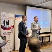 Zwei Männer stehen vor einer Leinwand, sie eröffnen eine Veranstaltung.